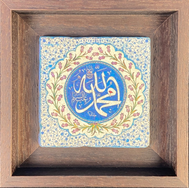 May include: Square ceramic tile with Arabic calligraphy in a blue circle, surrounded by a floral design. The tile is set in a dark brown wooden frame. The background has a light blue and white floral pattern.