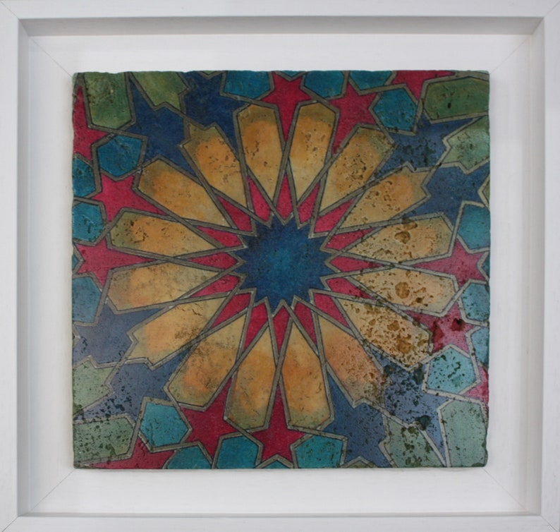 May include: A framed ceramic tile with a geometric pattern in shades of blue, red, gold, and green. The design features a central starburst with radiating lines and intricate details.