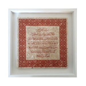 May include: A framed artwork with a white frame and a beige background. The artwork features a red border with a repeating pattern and Arabic calligraphy in red ink.
