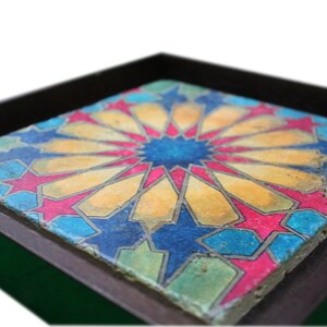 May include: A framed tile with a colorful geometric design. The tile features a central starburst pattern in shades of blue, yellow, and pink, surrounded by a repeating pattern of stars and geometric shapes.
