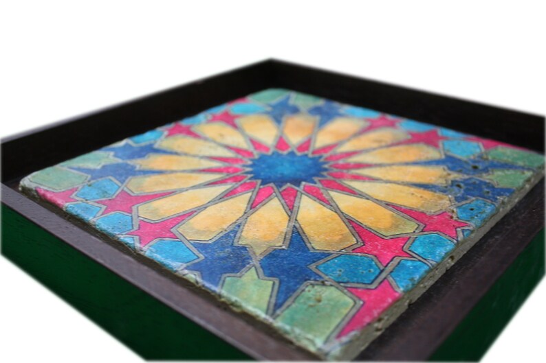 May include: A framed tile with a colorful geometric design. The tile features a central starburst pattern in shades of blue, yellow, and pink, surrounded by a repeating pattern of stars and geometric shapes.