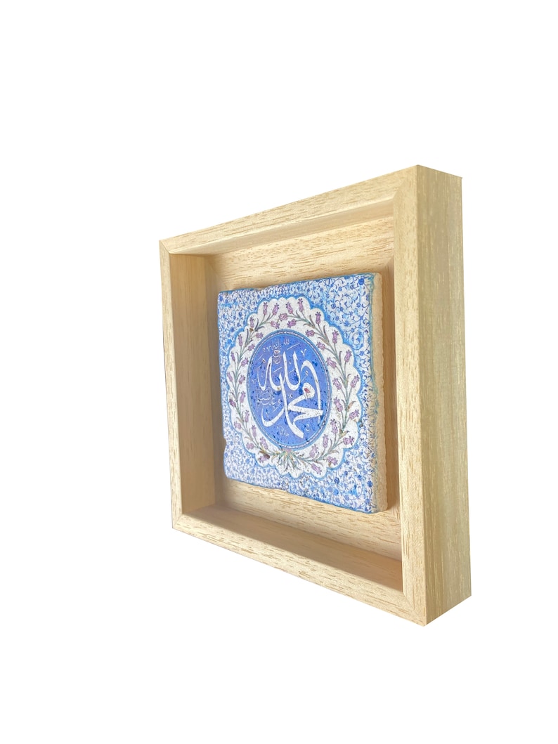 May include: A framed, square tile with Arabic calligraphy. The tile features a blue and white floral design with a central circular element. The frame is made of light-colored wood, creating a contrast with the tile's colors.