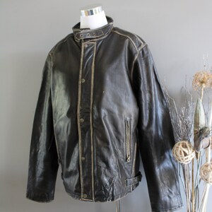 K&ouml;nnte beinhalten: Eine braune Lederjacke mit Stehkragen, Rei&szlig;verschluss und einem G&uuml;rtel in der Taille. Die Jacke hat einen Used-Look mit hellbraunen N&auml;hten.