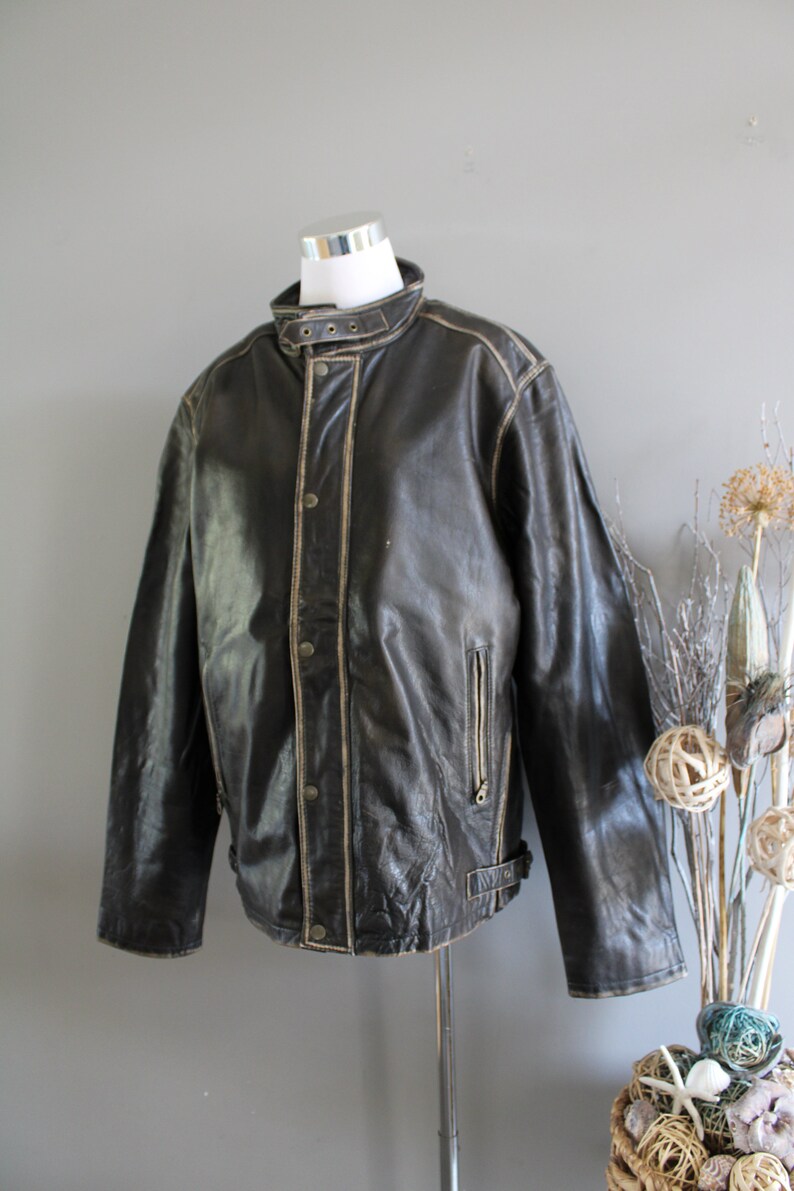K&ouml;nnte beinhalten: Eine braune Lederjacke mit Stehkragen, Rei&szlig;verschluss und einem G&uuml;rtel in der Taille. Die Jacke hat einen Used-Look mit hellbraunen N&auml;hten.