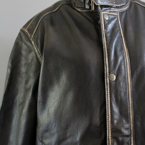 K&ouml;nnte beinhalten: Eine braune Lederjacke mit wei&szlig;em Steppsaum. Die Jacke hat einen Rei&szlig;verschluss und eine Knopfleiste.