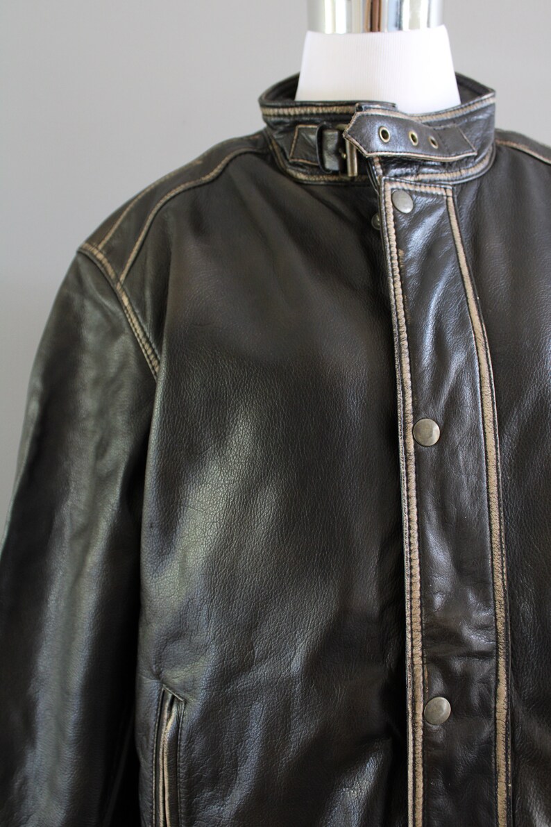 K&ouml;nnte beinhalten: Eine braune Lederjacke mit wei&szlig;em Steppsaum. Die Jacke hat einen Rei&szlig;verschluss und eine Knopfleiste.
