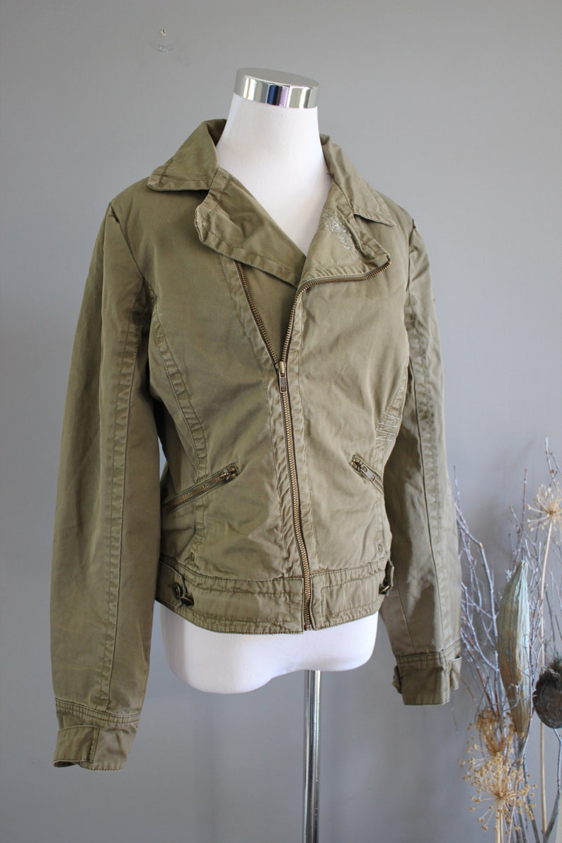 May include: A green cotton jacket with a zipper closure and multiple pockets. The jacket has a slightly fitted silhouette and a collar.