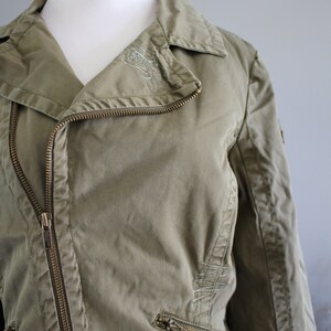 May include: A green cotton jacket with a zipper closure and a silver embroidered design on the collar. The jacket has two zippered pockets on the front and a zippered pocket on the left sleeve.