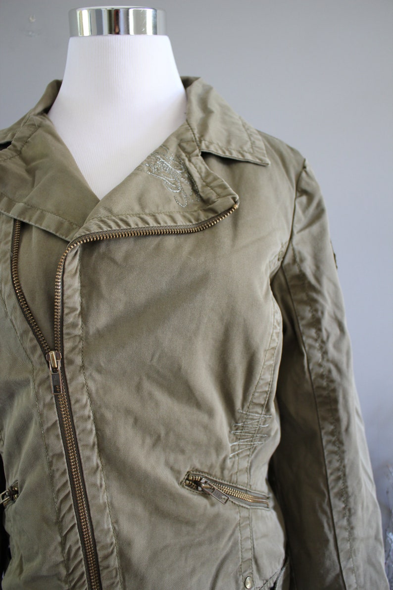 May include: A green cotton jacket with a zipper closure and a silver embroidered design on the collar. The jacket has two zippered pockets on the front and a zippered pocket on the left sleeve.
