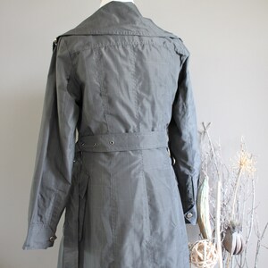 May include: A gray, belted trench coat with a collar and long sleeves. The coat has a checkered pattern and a belt with silver buckles.