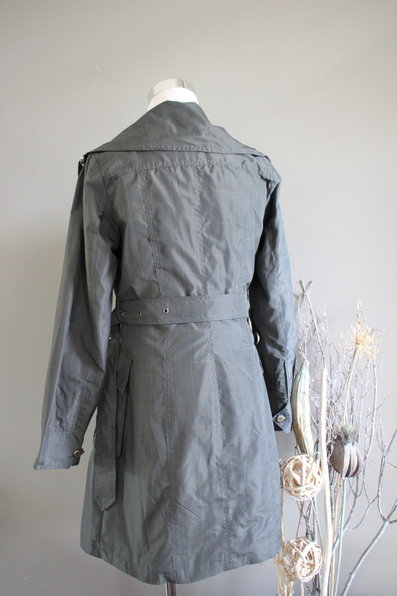 May include: A gray, belted trench coat with a collar and long sleeves. The coat has a checkered pattern and a belt with silver buckles.