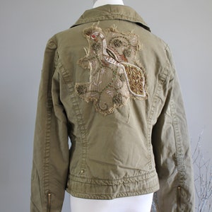 May include: A green utility jacket with a large embroidered design on the back. The design features a bird with gold and brown accents.
