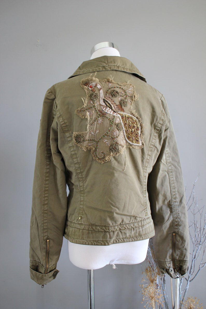 May include: A green utility jacket with a large embroidered design on the back. The design features a bird with gold and brown accents.