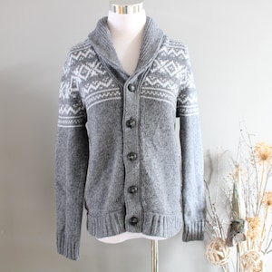 May include: Grey cardigan sweater with a white and grey geometric pattern on the collar and cuffs. The sweater has a shawl collar and a button front.