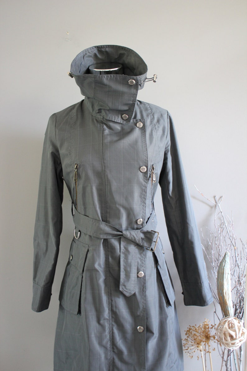 May include: A gray, belted trench coat with a high collar and a button closure. The coat has a pinstripe pattern and a zippered pocket on the left side.