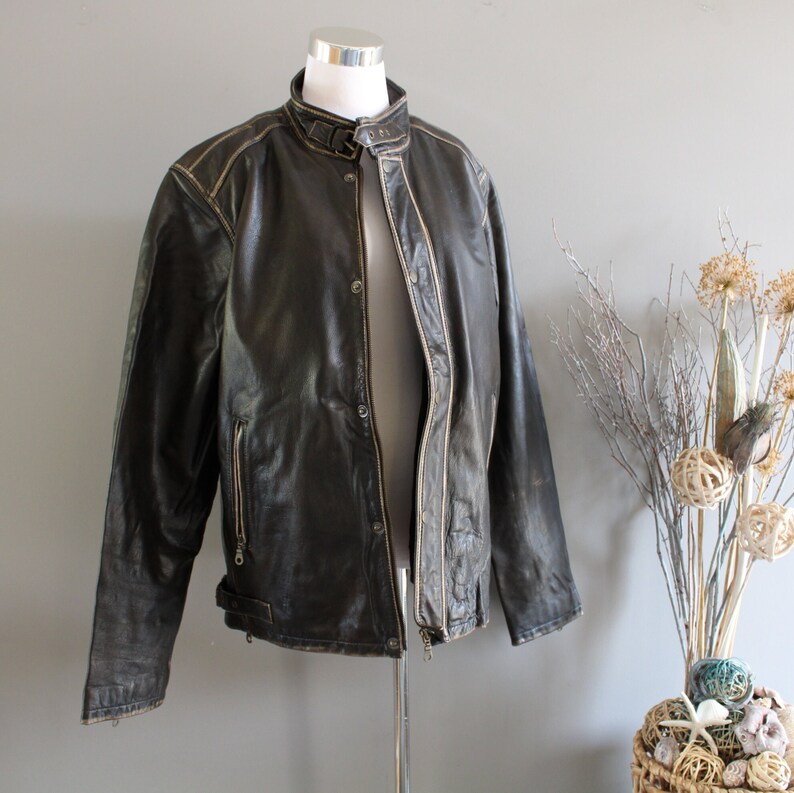 K&ouml;nnte beinhalten: Eine braune Lederjacke im Motorrad-Stil mit Rei&szlig;verschluss und Druckknopfleiste. Die Jacke hat einen Used-Look mit hellbraunen N&auml;hten.