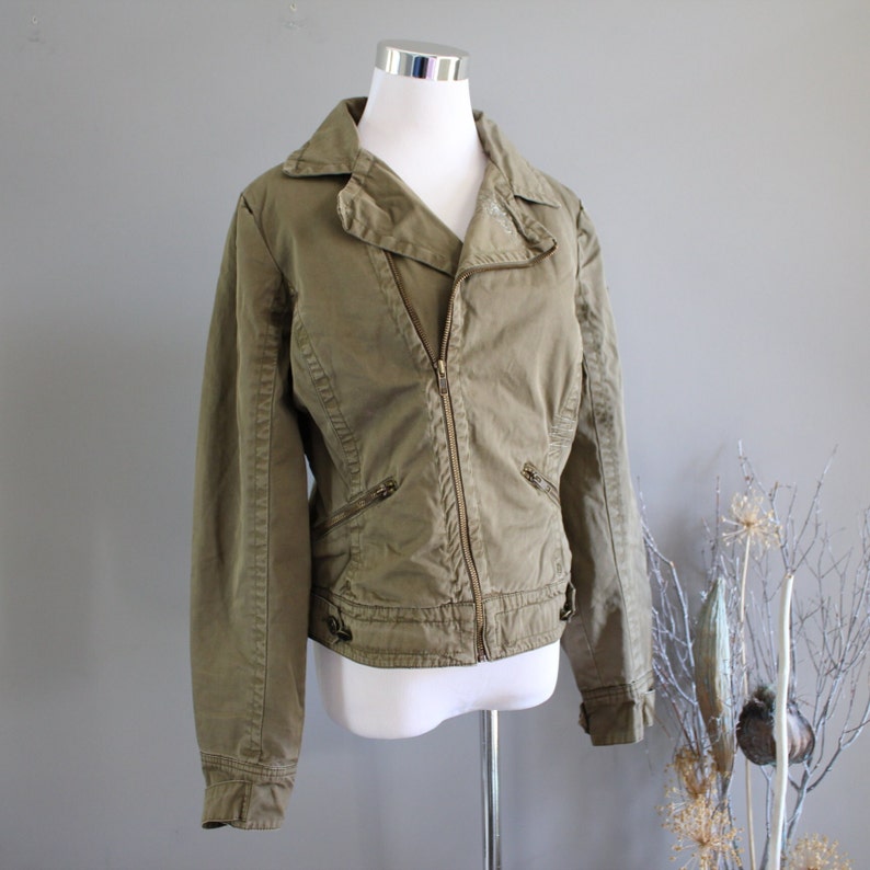May include: A green, zippered jacket with a collar and long sleeves. The jacket has a fitted silhouette and a slightly asymmetrical zipper closure.