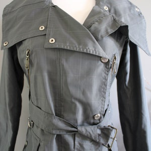May include: A gray plaid trench coat with a belted waist and a zipper closure. The coat has a collar that can be folded up and fastened with a button.