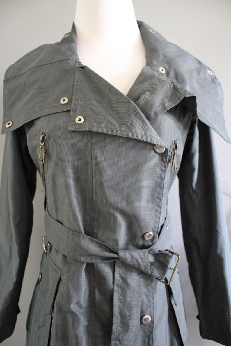 May include: A gray plaid trench coat with a belted waist and a zipper closure. The coat has a collar that can be folded up and fastened with a button.