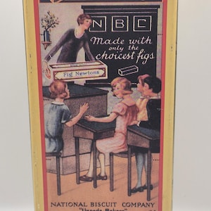 May include: Vintage Fig Newtons tin with a yellow and red design. The tin features illustrations of the product and a school scene. Text includes "Fig Newtons" and "Made with only the choicest figs."