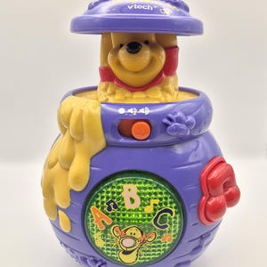 May include: A purple and yellow VTech Winnie the Pooh toy. The toy features a smiling Winnie the Pooh character, a honey pot design, and an ABC and music button. The toy has an orange button and the VTech logo.