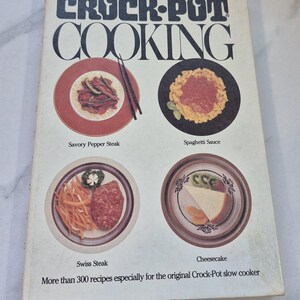 May include: Vintage "Crock-Pot Cooking" cookbook with images of prepared dishes. The cover features the title in bold black letters and images of Savory Pepper Steak, Spaghetti Sauce, Swiss Steak, and Cheesecake. The book promises over 300 recipes for the Crock-Pot slow cooker.
