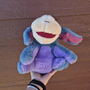 May include: A plush toy character featuring a soft, light beige muzzle, a wide dark red mouth, and long, floppy ears with purple inner linings. The body is covered in textured blue and purple fur, with a rounded shape and soft, plush limbs.