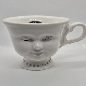May include: A white ceramic teacup with a face design. The cup features a winking eye, an open eye with blue iris, a nose, and a smiling mouth. The base of the cup has a dotted pattern. The handle is on the right side.
