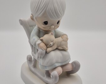 May include: A porcelain figurine of a seated elderly person in a rocking chair, wearing glasses and holding a sleeping cat. The figurine is primarily white, with pastel blue and pink accents. The base is oval-shaped.