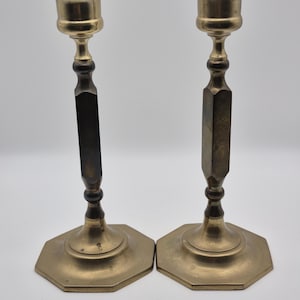 May include: A pair of matching brass candlesticks featuring a flared top cup, a slender central stem with a faceted hexagonal middle section, and a wide, flat octagonal base. The metal has a polished finish with some natural patina and dark tones.