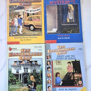 May include: Four vintage paperback books from "The Baby-Sitters Club" series. The covers feature illustrations of children and houses, with titles like "Good-bye Stacey, Good-bye" and "Kristy and the Secret of Susan". The books are by Ann M. Martin.