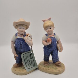 May include: Two porcelain figurines of children wearing blue denim overalls and white shirts. One figure wears a tan cowboy hat and carries a book, while the other has a pink hair bow and holds an orange fruit. A tag reads DENIM DAYS BY HOMCO.