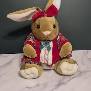 May include: A plush brown bunny with long ears and a red bow. The bunny wears a red floral print dress with a green bow and white trim. The bunny is sitting on a marble surface.