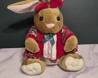 May include: A plush brown bunny with long ears and a red bow. The bunny wears a red floral print dress with a green bow and white trim. The bunny is sitting on a marble surface.