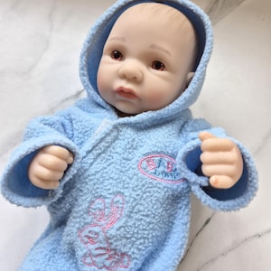 May include: A lifelike baby doll wearing a light blue hooded robe. The robe has a pink embroidered bunny and the text "BABY BORN". The doll has realistic features, including open eyes and a soft complexion. The doll's hands are in a fist.