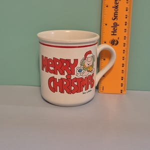 May include: White ceramic Christmas mug with red trim and lettering. The mug features the words "MERRY CHRISTMAS" in red, along with a cartoon illustration of a character wearing a Santa hat. The mug is approximately 4 inches tall.