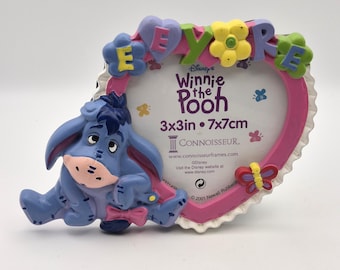 May include: A heart-shaped picture frame featuring a blue Eeyore character on the left side. The frame is pink with a white scalloped edge and decorated with colorful flowers and letters spelling EEYORE. The center insert reads Winnie the Pooh 3x3in.