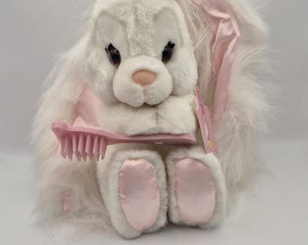 May include: A plush white rabbit toy with long, fluffy fur, featuring a pink satin bow on its head and matching pink satin fabric on its feet. The rabbit holds a small pink plastic hairbrush in its paws and has a pink tag attached to its side.