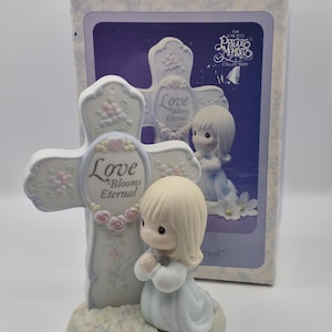 May include: A Precious Moments figurine of a kneeling girl in a light blue dress, praying before a cross. The cross is adorned with the words "Love Blooms Eternal" and floral accents. The figurine is on a base with a grassy texture, and the box is in the background.