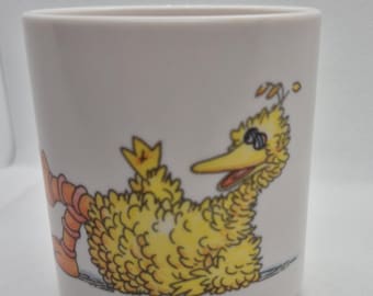 May include: A white ceramic mug featuring a cartoon illustration of Big Bird from Sesame Street. The yellow bird is depicted with sunglasses, orange legs, and a long yellow beak. The mug is cylindrical and has a simple, clean design.