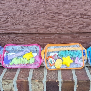 May include: Four rectangular clear plastic pouches with colored zippers and trim in purple, pink, orange, and blue. Each pouch contains a variety of small colorful items, including yellow star-shaped objects and textured green coils.