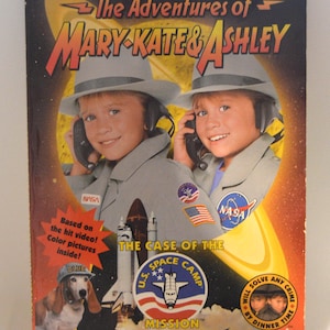 May include: A book cover for "The Adventures of Mary-Kate & Ashley: The Case of the Space Camp Mission." The cover features two girls in gray NASA uniforms and hats, with a dog wearing a hat. The book has a red and yellow background.