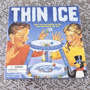 May include: A board game box titled "THIN ICE" with a blue background and illustrations of children and a penguin. The game features a marble-dropping mechanism. The box includes a warning about choking hazards and is for ages 5 and up.