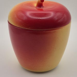 May include: A ceramic apple-shaped container with a lid. The container is painted with a gradient of colors, transitioning from red at the top to yellow at the bottom. The lid has a stem-like handle.