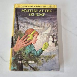 May include: A vintage book cover for "Mystery at the Ski Jump" from the Nancy Drew Mystery Stories series. The cover features an illustration of a young woman in winter attire, a ski jump, and the text "by Carolyn Keene".