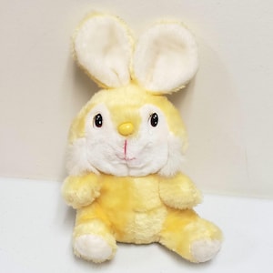 May include: A plush yellow bunny rabbit with large, upright ears. The bunny has a white face, chest, and inner ears, with black eyes and a red stitched mouth. The bunny is sitting upright, with its arms and legs extended.