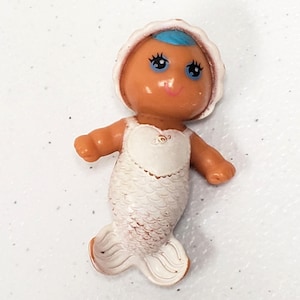 May include: A small plastic mermaid figurine with tan skin, bright blue hair, and a white scalloped bonnet. The mermaid has a white bodice with a heart-shaped chest detail and a white fish tail with embossed scale patterns and a flared fin base.
