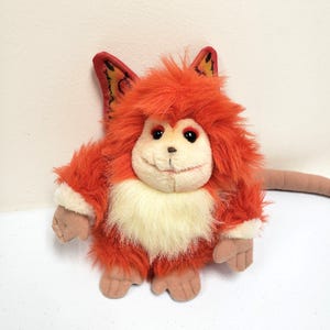 May include: A plush toy creature with orange fur, red ears, and a cream-colored chest. The toy has a smiling face with red eyes and a long, brown tail. The toy is a stuffed animal.