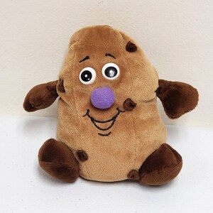May include: A plush toy shaped like a brown cookie with dark brown chocolate chips. The toy features two large plastic googly eyes, a round purple fabric nose, and a black embroidered smiling mouth. It has two brown arms and two brown feet.