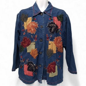 May include: A blue denim jacket featuring a button-down front and long sleeves. The jacket is decorated with embroidered autumn leaf motifs in shades of orange, red, yellow, and dark green, interspersed with small square textured patches.